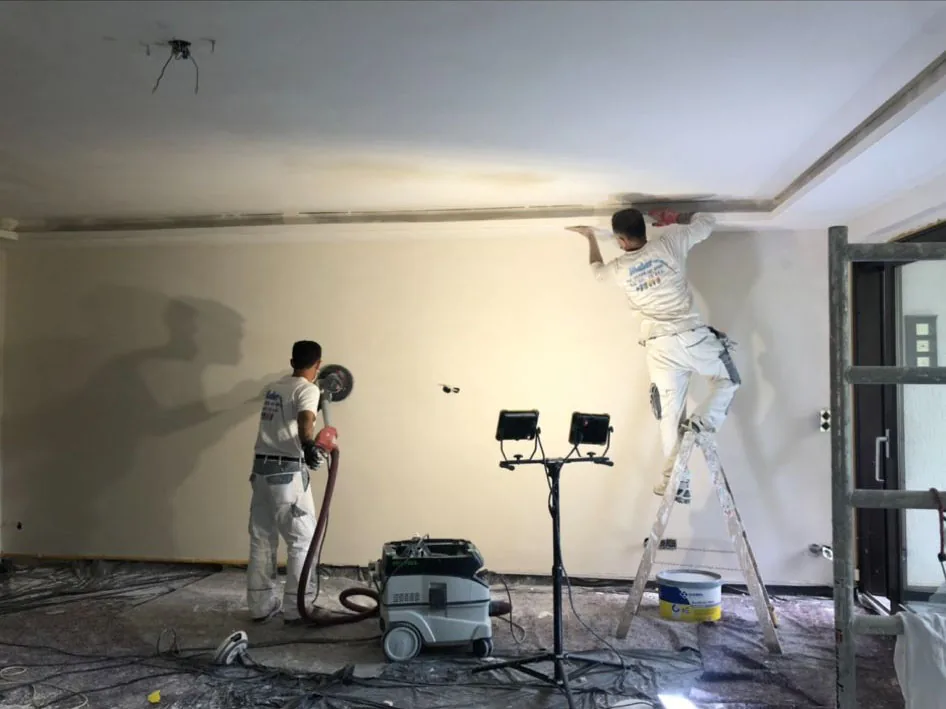 Zwei Maler von HAMA schleifen und spachteln Wände und Decke eines Wohnraums.